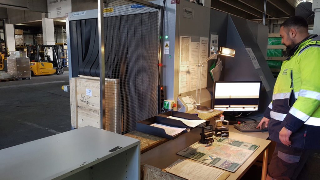 Ein Mitarbeiter bedient per PC eine Röntgenanlage in der gerade ein größeres Frachtgut hereingefahren wird, im hintergrund eine große Lagerhalle